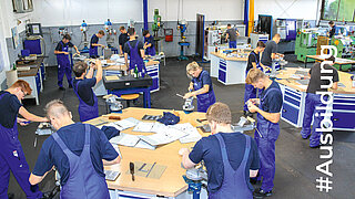 Auszubildende in blauen Overalls stehen im Kreis um Werkbänke und bearbeiten Bauteile