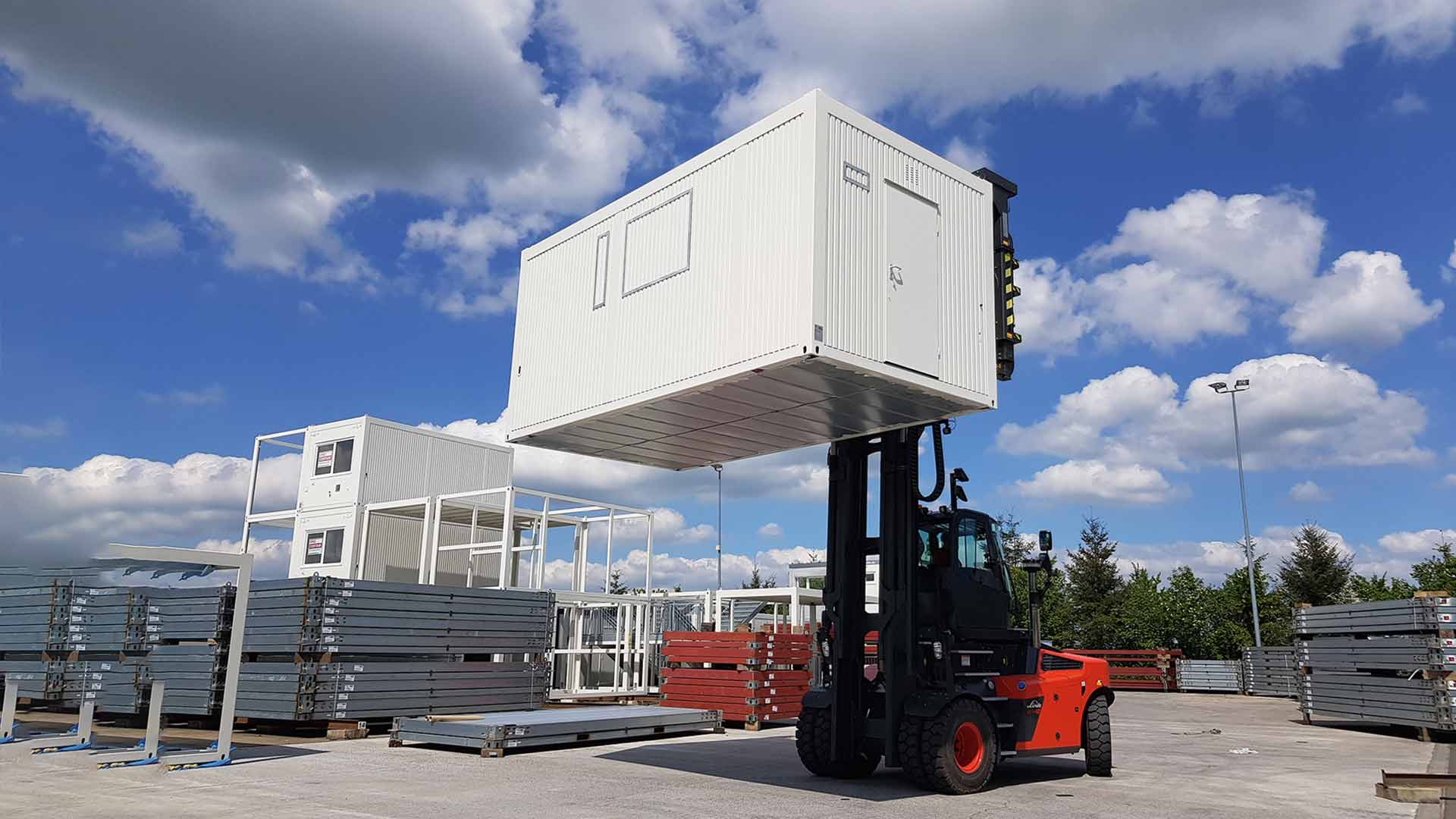 A red forklift truck lifts a white container high up with the help of a wide spreader