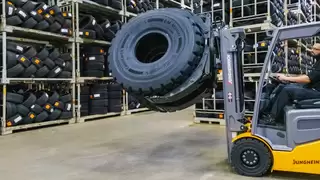 A forklift truck with rotating attachment lifts and transports two tyres through a warehouse