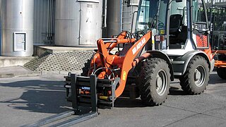 A construction machine, equipped with a forklift fork as attachment, stands in front of silos