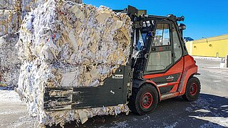 Eine rote Baumaschine transportiert einen gepressten Ballen Altpapier in einer Ballenklammer