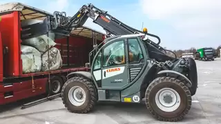 A construction machine unloads a red truck with a bale clamp attached to the crane