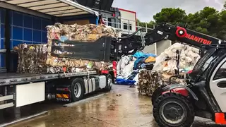 A forklift truck with a bale clamp lifts a cube of pressed waste paper onto a truck bed