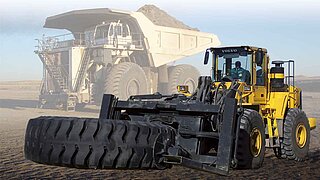 A yellow construction machine with a wide fork transports a large tyre on a sandy surface