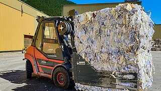 Un chariot élévateur rouge tient un grand cube de vieux papiers compactés dans une pince à balles