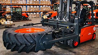 A red construction machine transports a large tyre horizontally with a bale clamp