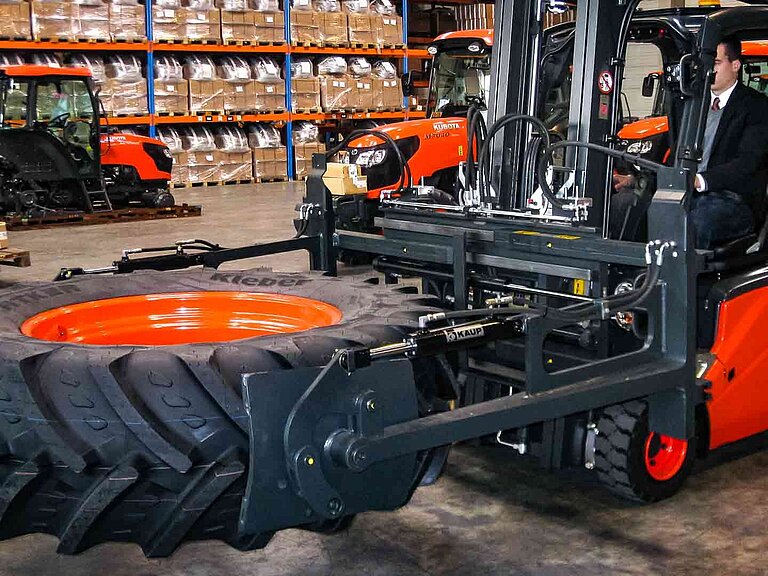 Un engin de chantier rouge transporte un grand pneu horizontalement à l'aide d'une pince à balles