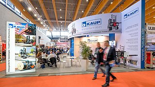 Vista de un stand de feria de KAUP con dos personas pasando en primer plano