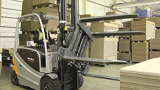 A silver forklift truck with rotating attachment loads a stack of boxes in a warehouse