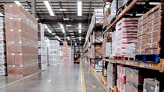 A forklift truck drives along an aisle with filled storage racks and stacks of boxes