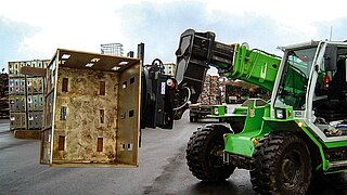 Eine grüne Baumaschine dreht eine angehobene Holzkiste mithilfe eines rotierenden Anbaugeräts