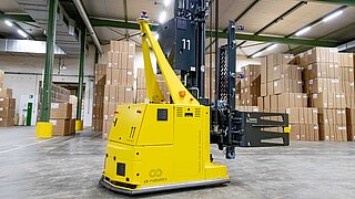 A driverless, yellow transport system with bale clamp in front of a large number of stacked cartons