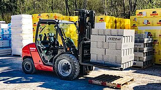 A red forklift truck transports stacked bricks without a pallet across an open storage area