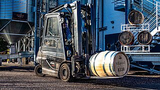 A black forklift truck transports a wooden spirit barrel across a factory site
