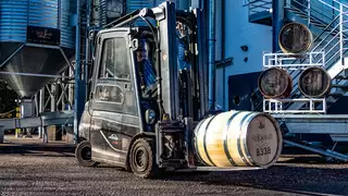 A black forklift truck transports a wooden spirit barrel across a factory site