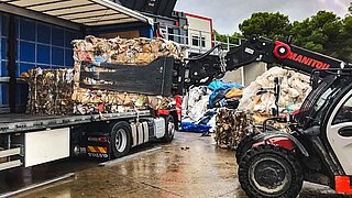 A forklift truck with a bale clamp lifts a cube of pressed waste paper onto a truck bed