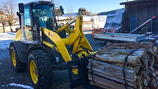 Eine gelbe Baumaschine nimmt mehrere Rollen aus Holzscheiten mit einem Zinkenverstellgerät auf