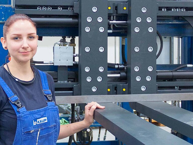 Junge, lächelnde Frau in blauem KAUP-Overall mit einer Hand auf der Gabel eines Staplers