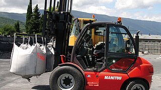 A construction machine transports several white bags hanging from the forks of the forklift truck