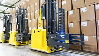 Dos carretillas elevadoras amarillas sin conductor delante de un número de cajas de cartón apiladas