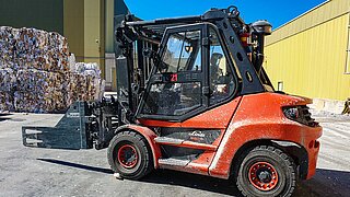 Une machine de chantier rouge avec pince à balles devant des balles de vieux papier pressées