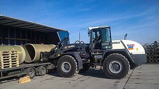 A white construction machine loads a pipe onto the fork for transport from a truck bed