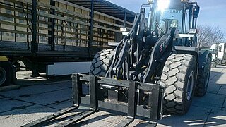 A construction machine with mounted sideshift is parked in front of a lorry with open loading area