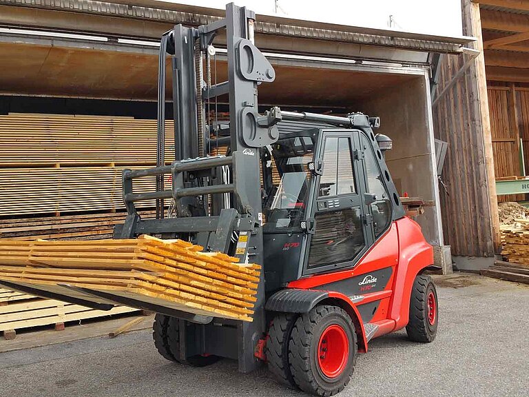 A red forklift truck transports several stacked wooden boards on a two-pronged attachment