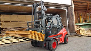 A red forklift truck transports several stacked wooden boards on a two-pronged attachment