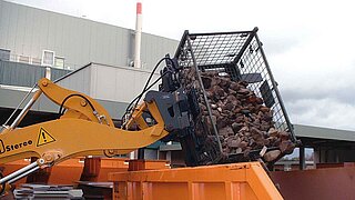 Una máquina de construcción vierte una caja de escombros en un contenedor de construcción naranja