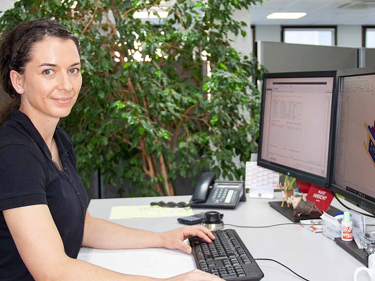 Une femme est assise à son bureau devant deux écrans et regarde la caméra par-dessus son épaule