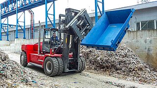 Eine rote Baumaschine mit drehbarem Anbaugerät rotiert einen blauen Bauschuttbehälter