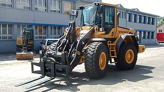 Gros engin de chantier, équipé d'une fourche de chariot élévateur, devant un immeuble de bureaux