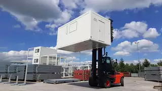 A red forklift truck lifts a white container high up with the help of a wide spreader