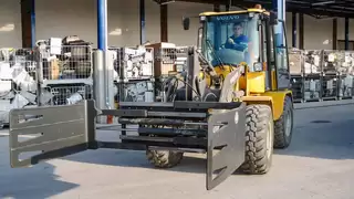 A yellow construction machine with attached wide clamp in front of boxes of construction waste