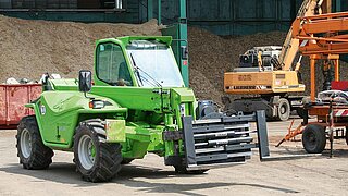 A construction machine with mounted forklift fork in front of sand mountains and other machines