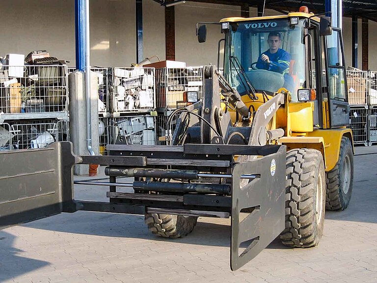 A yellow construction machine with attached wide clamp in front of boxes of construction waste