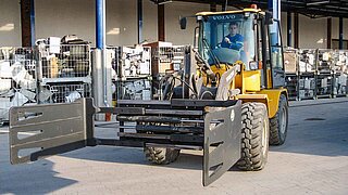 A yellow construction machine with attached wide clamp in front of boxes of construction waste