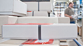 Elongated white parcels with red stickers are stacked on top of each other in a warehouse