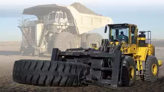 A yellow construction machine with a wide fork transports a large tyre on a sandy surface