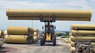 A large construction machine lifts two long pipes while passing through a narrow passageway