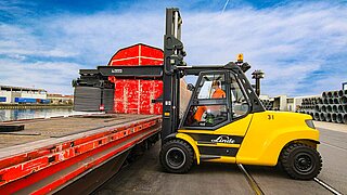 A yellow construction machine places a large roll of wire on the loading area of a train wagon