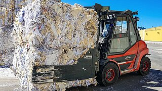 Un chariot élévateur rouge transporte deux grandes balles compressées de vieux papier avec une pince