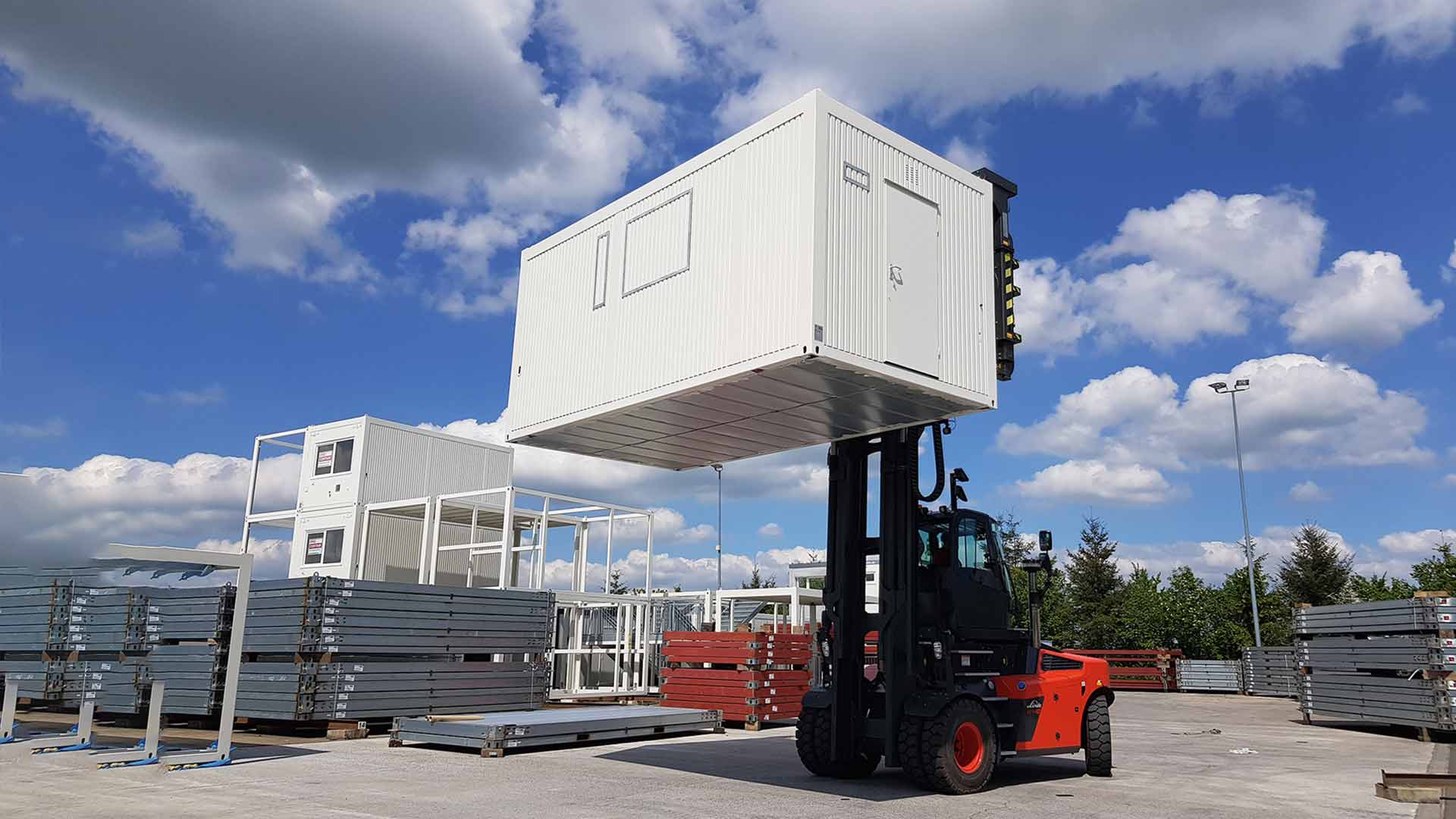 Ein roter Gabelstapler hebt einen weißen Container weit nach oben mithilfe eines breiten Spreaders