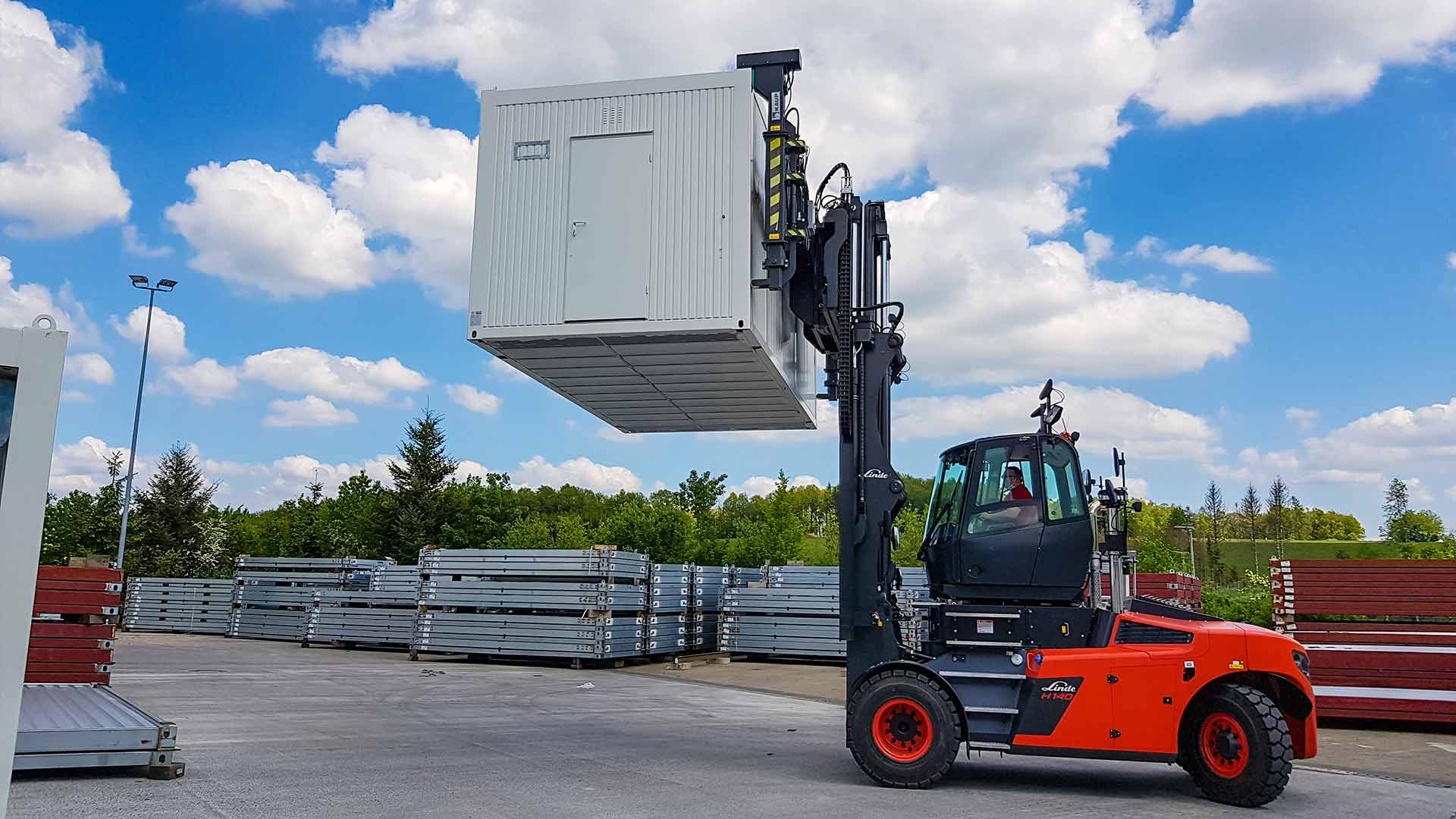 Eine rote Baumaschine transportiert einen silbernen Container mit Tür über einen Lagerplatz