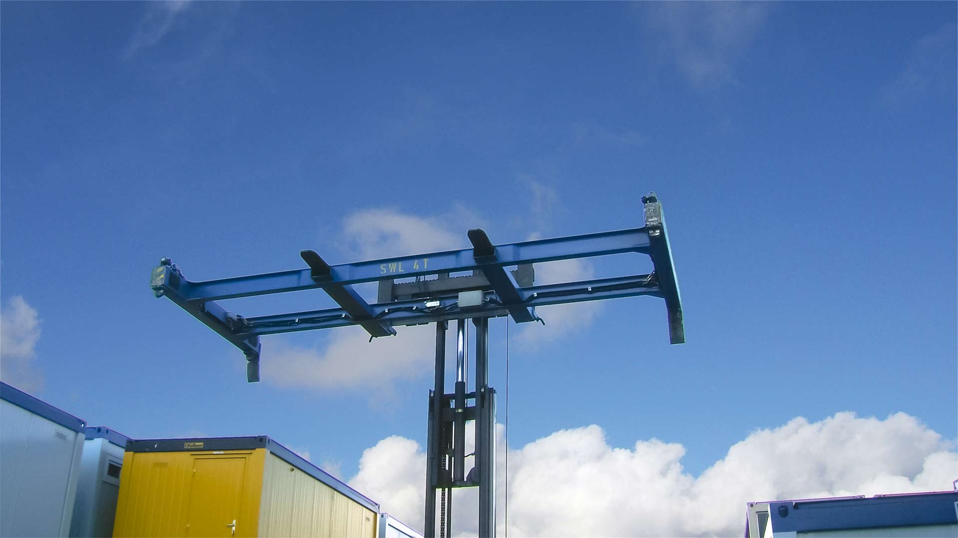 A blue container spreader mounted on a forklift truck is extended far upwards