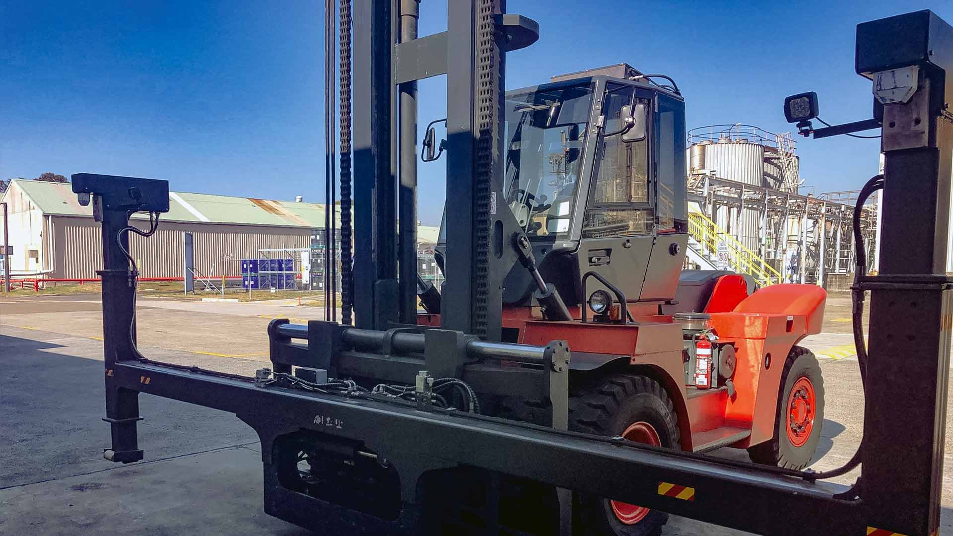 Red forklift truck with wide attachment in front of silos and an industrial plant in the background