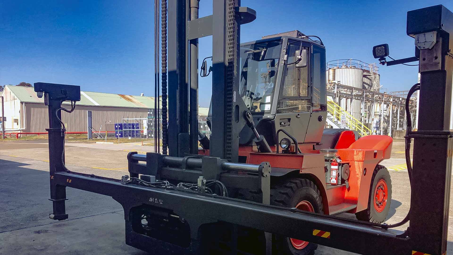 Carretilla elevadora roja con accesorio ancho delante de silos y una planta industrial al fondo