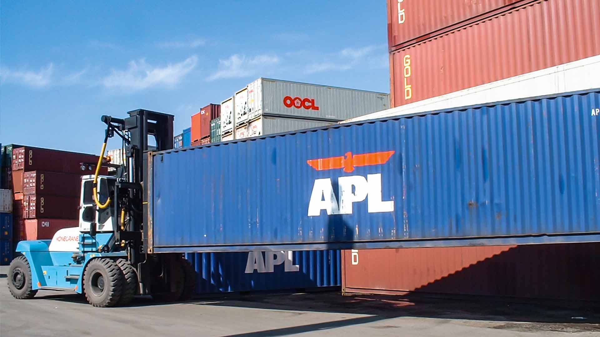 A forklift truck equipped with a front spreader transports a blue container