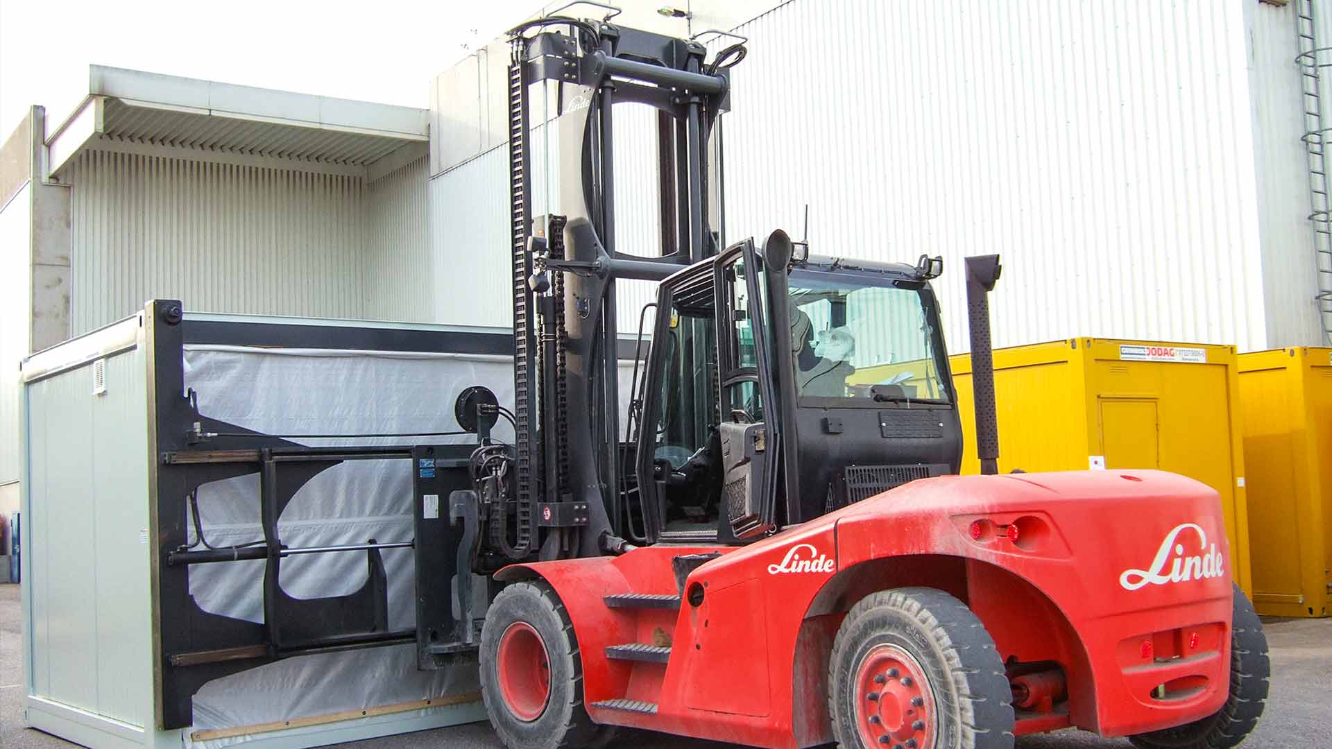 Gabelstapler mit breitem Anbaugerät, der einen Container an seiner Längsseite zum Transport aufnimmt